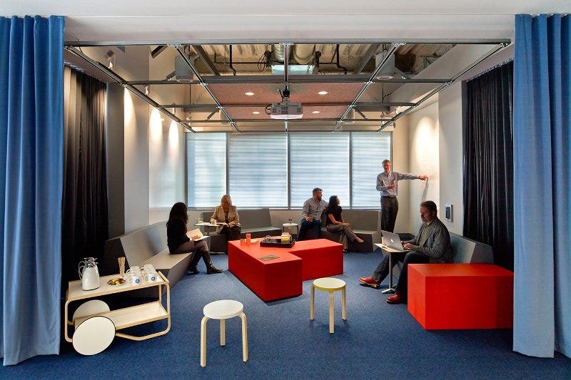 Espace lounge de bureau, des collaborateurs échangent autour de tables cubiques rouges pendant qu’un homme présente au tableau