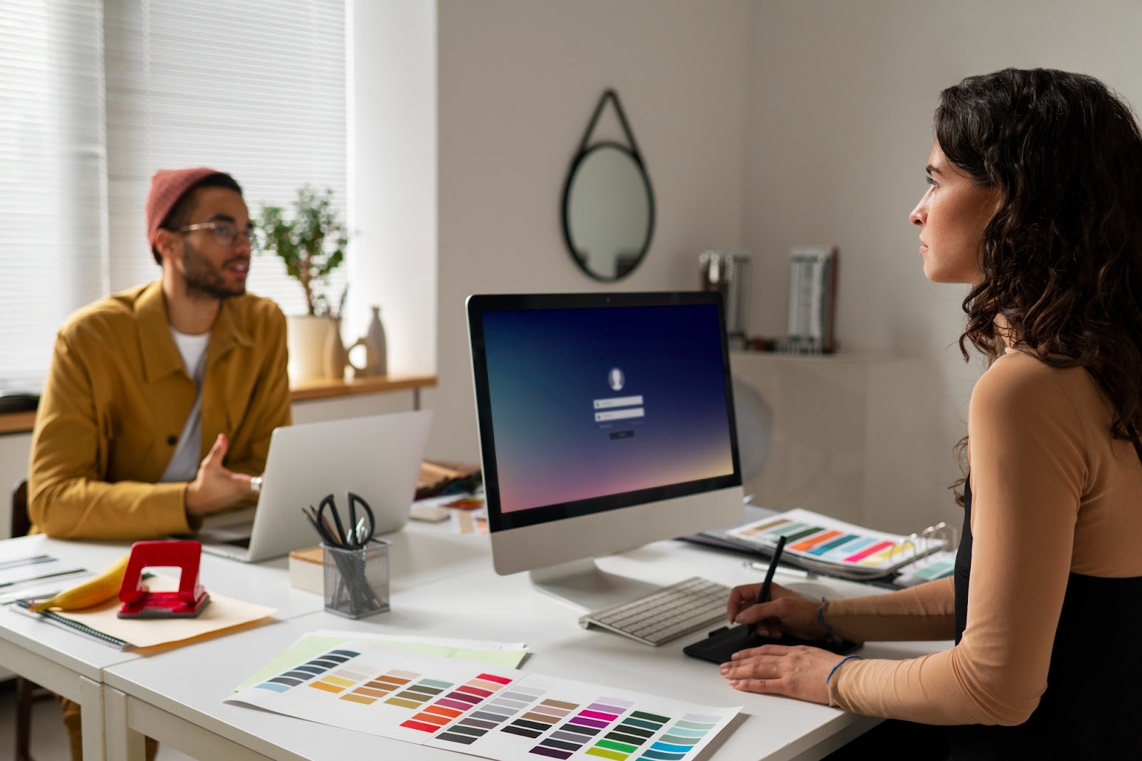 Deux designers échangent en studio devant un iMac, avec une palette de couleurs et une tablette graphique sur le bureau