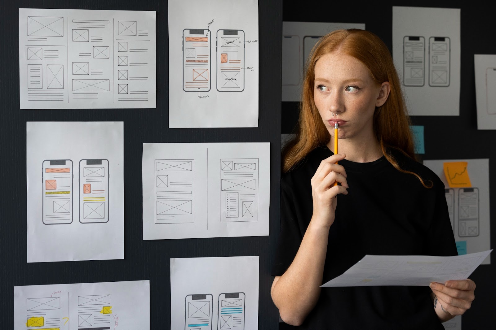Designer UX pensive devant un mur de wireframes papier d’écrans mobiles et web, un crayon à la main