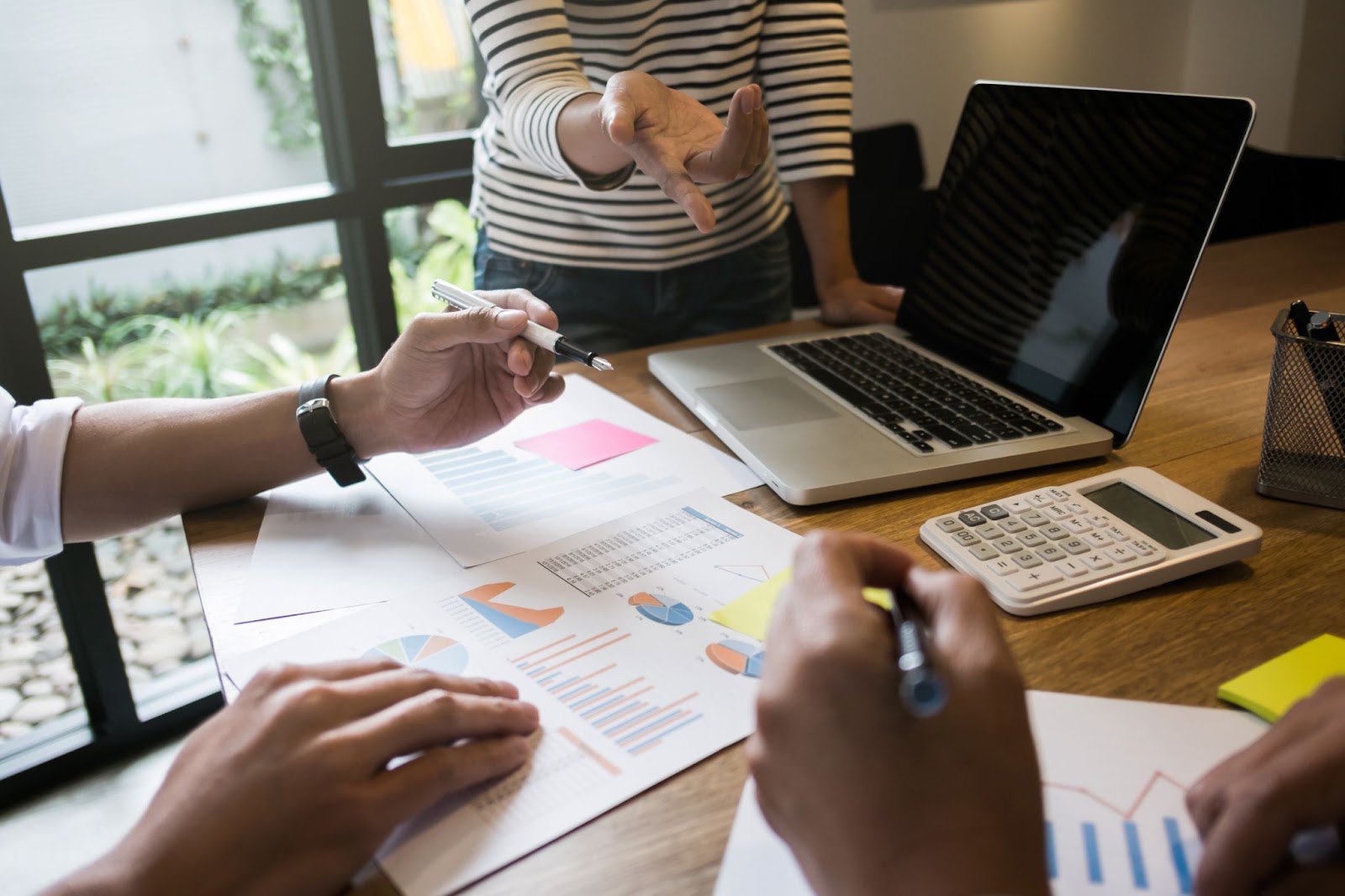 Réunion d’analyse, des mains pointent des graphiques imprimés sur un bureau à côté d’un ordinateur portable et d’une calculatrice