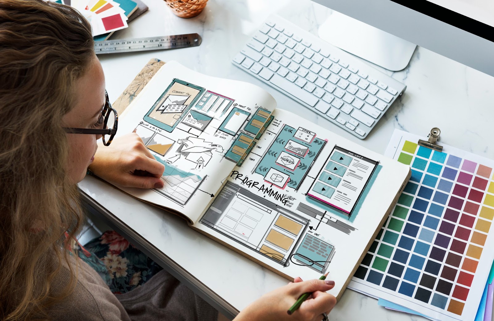 Designer à lunettes étudiant un carnet de croquis d’interfaces, près d’un clavier et d’un nuancier de couleurs